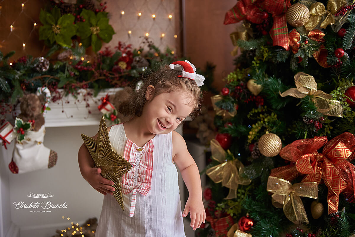 Menina com síndrome de down, sorrindo, feliz, segurando uma estrela dourada, em uma sala com decoração de natal, em Caxias do Sul, RS.