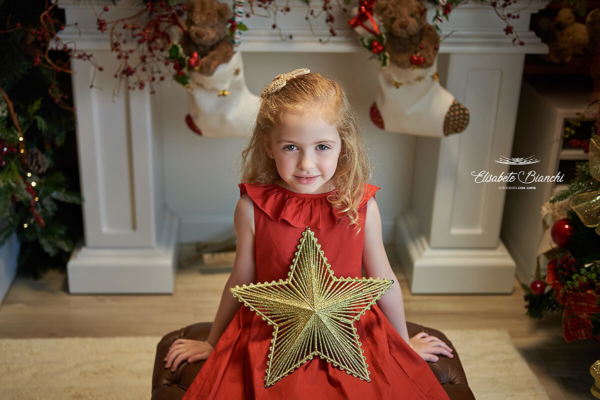 Menina com vestido vermelho, com uma estrela dourada no colo, sentada em frente à lareira, em Caxias do Sul, RS.