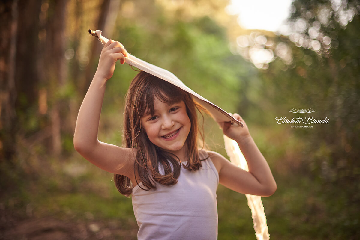 Menina sorrindo e segurando uma pandorga sobre  sua cabeça, para fazer sombra em seu rosto.