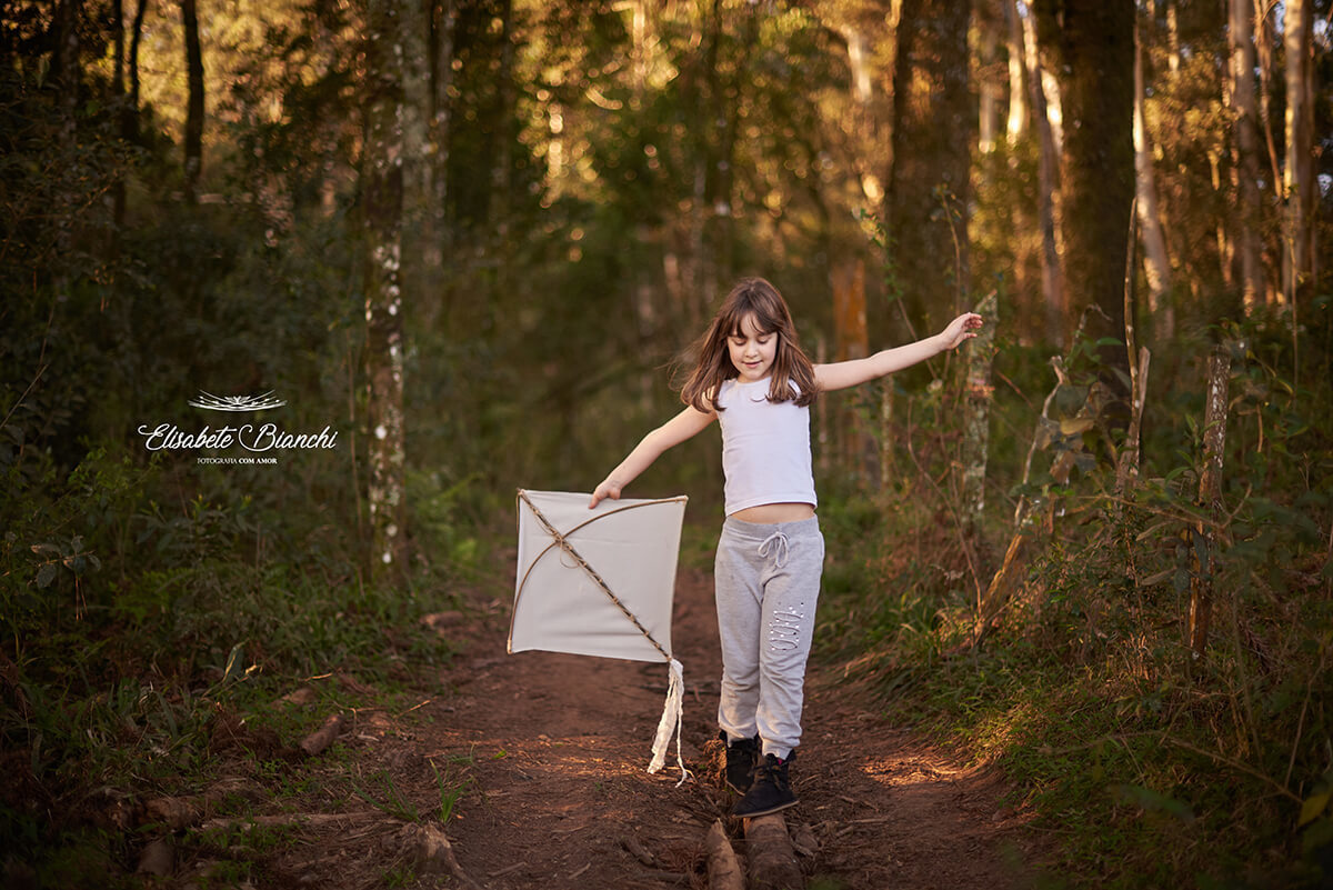 Menina se equilibrando com os braços abertos e uma pandorga na mão, sobre um tronco de árvore jogado no chão, no meio de uma floresta, em Caxias do Sul.