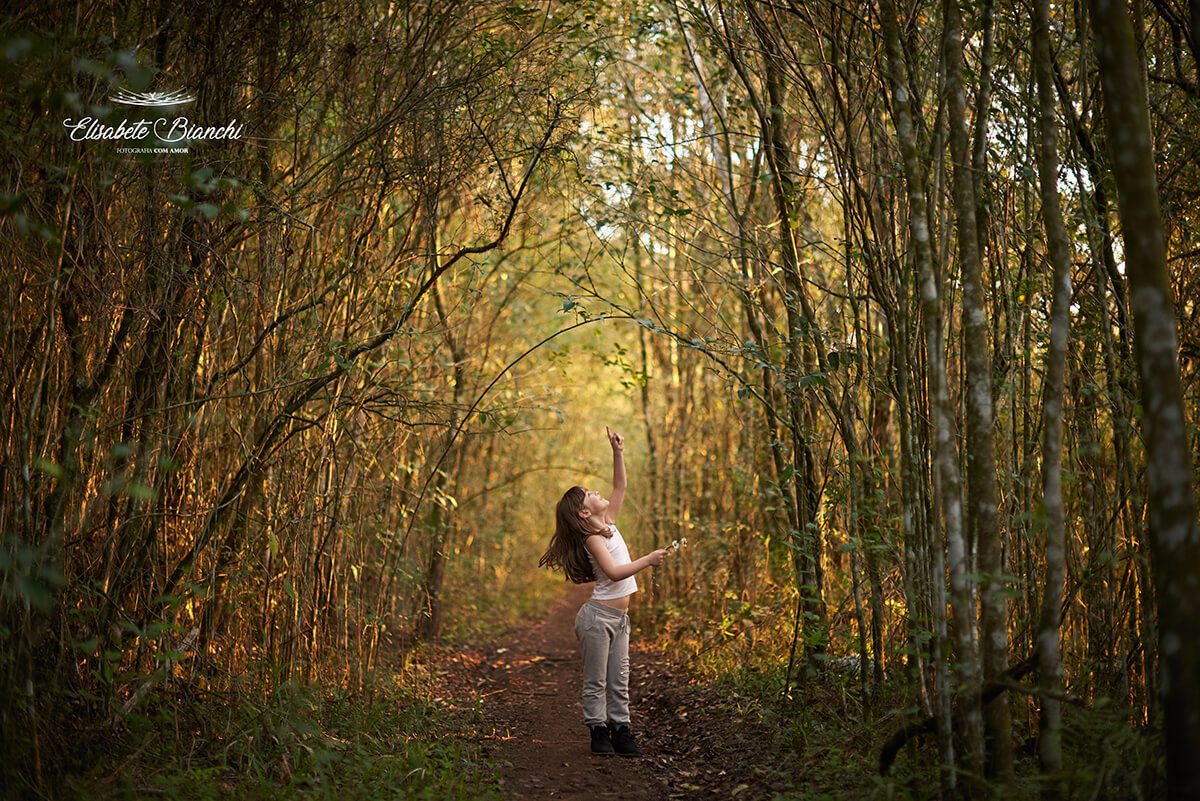 Menina apontando para o céu, no meio de árvores altas.