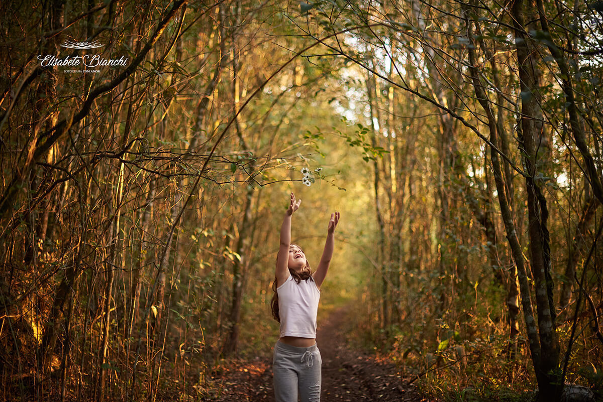 Menina apontando os braços levantados para o alto, em uma floresta.