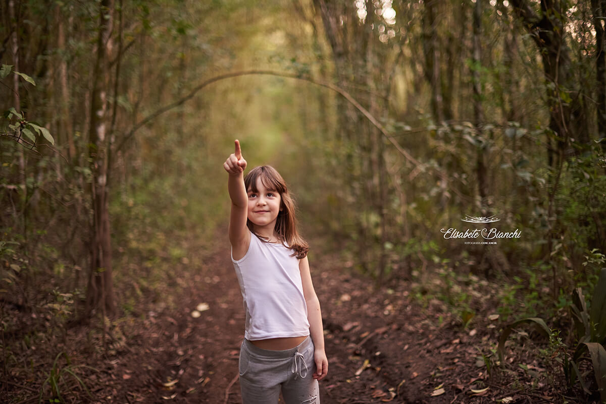 Menina apontando para o alto, como que mostrando algo que viu, em meio à floresta.