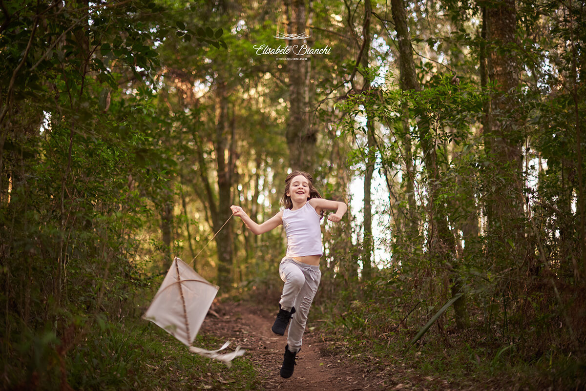 Menina em movimento no ar, com uma perna dobrada, como que correndo, com a corda de uma pandorga nas mãos, tentando empiná-la. Ela está em uma trilha na floresta.