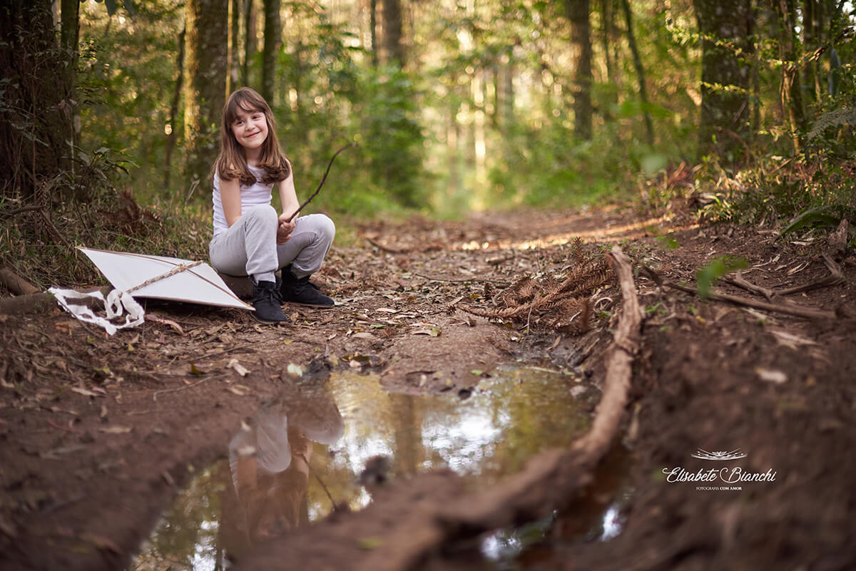 Menina sentada na beira de uma estrada de chão batido, próxima à uma poça de água, descansando, com sua pipa ao lado.