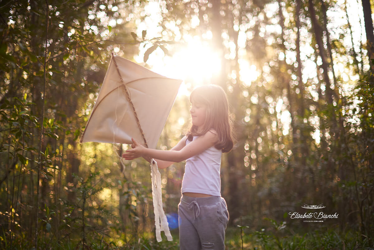 Menina arrumando sua pandorga, em meio à natureza. A luz do sol está acima de sua cabeça.