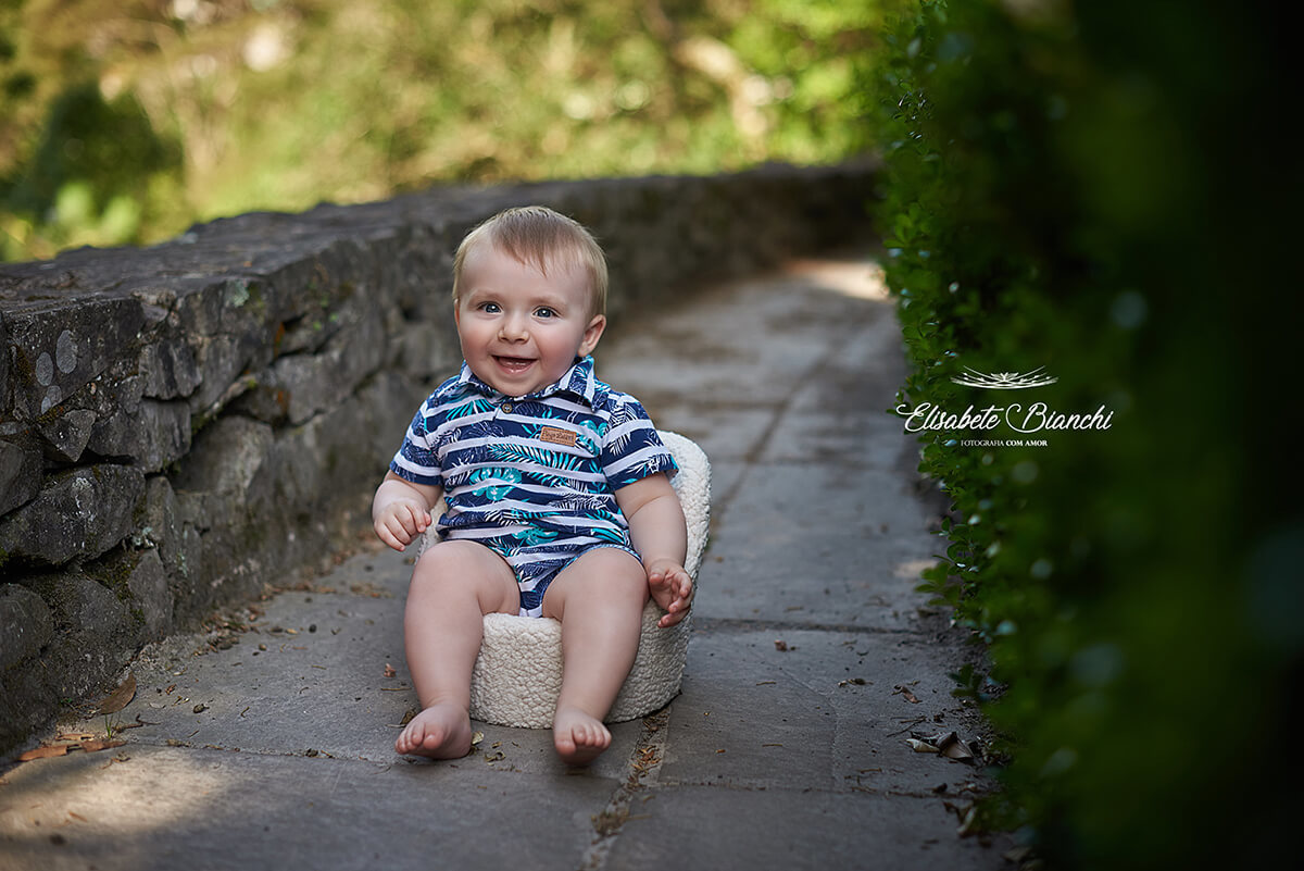 Bebê de 9 meses de idade, sentado em uma poltrona, em um caminho estreito, entre muro de pedras e uma parede verde.