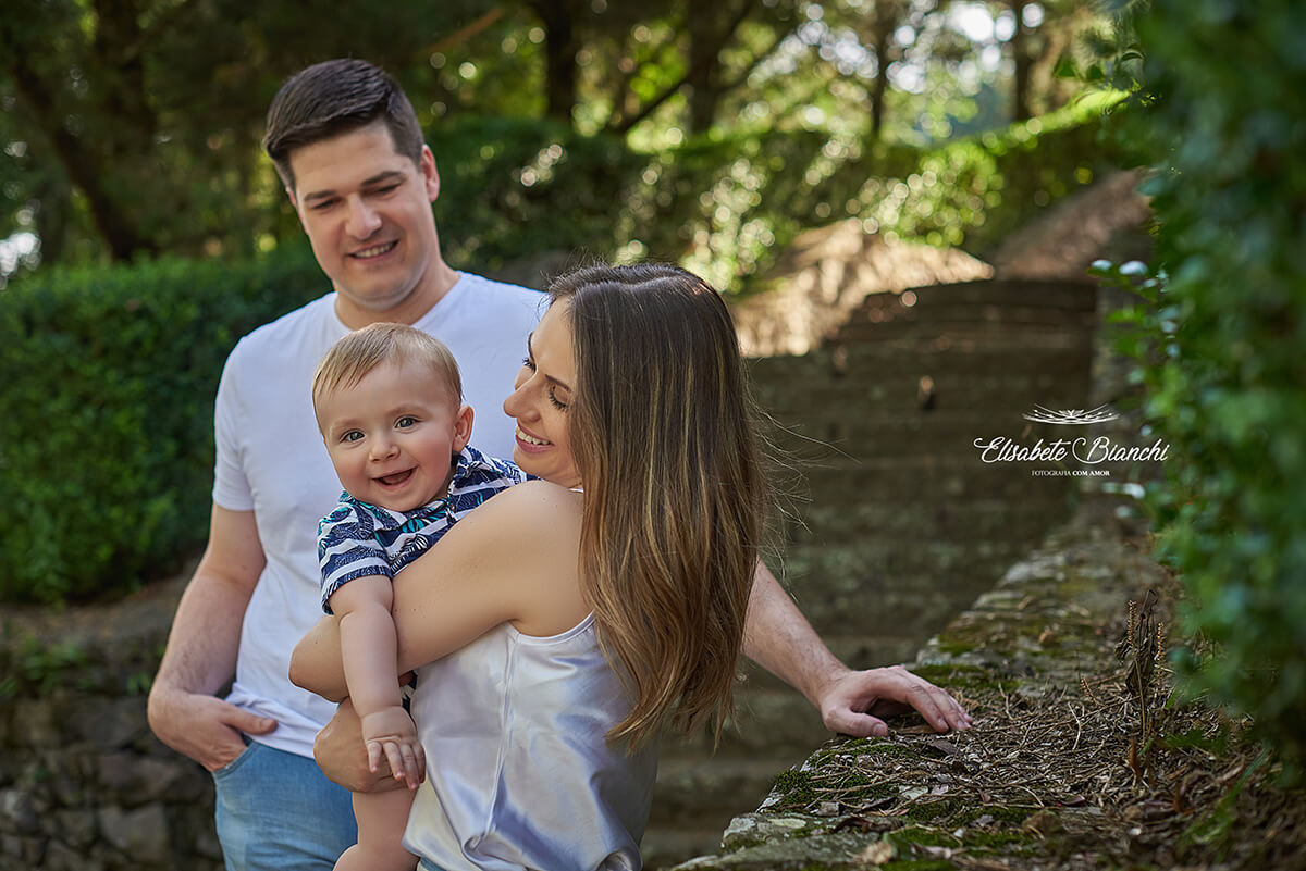 Mãe segura seu bebê no colo, olhando para a câmera. O pai está atrás deles, com uma mão no bolso. Todos sorriem.