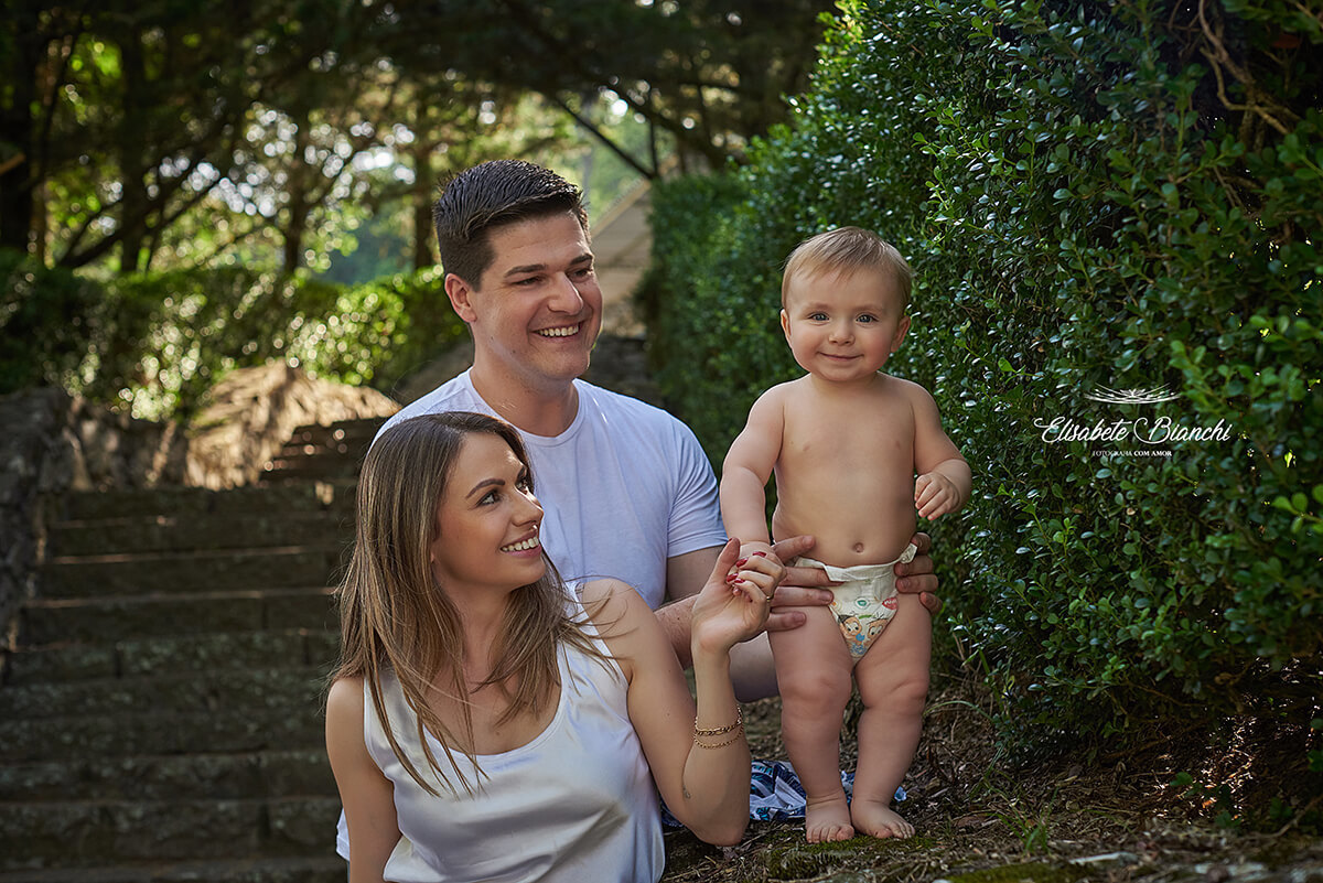 Pai e mãe seguram seu bebê de pé, só de fraldinhas, sobre um muro.