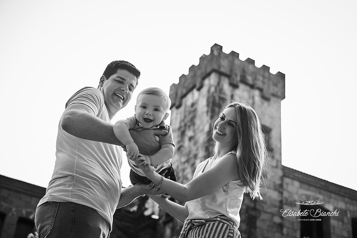 Pais brincando de balanço nos braços com seu bebê, vistos de baixo pra cima, em frente ao Castelo Chateau La Cave, em Caxias do Sul.
