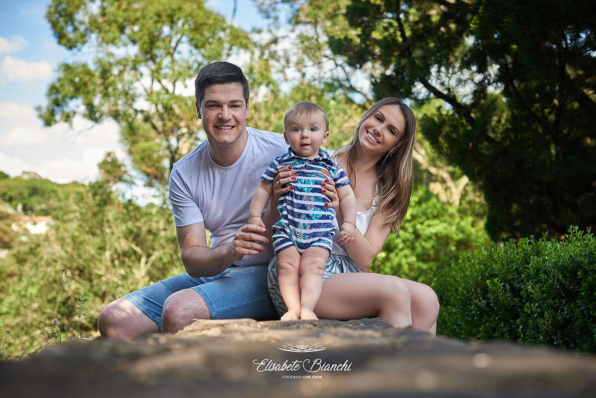Pai e mãe sentados em um muro, cada um com as pernas para um lado do muro, segurando seu bebê de pé sobre o muro. Os três olham para a câmera.