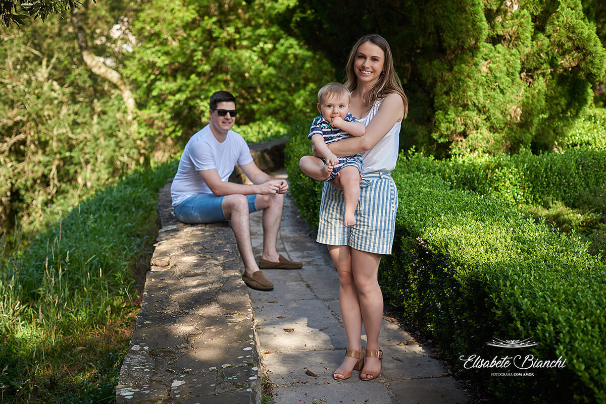 Mãe, de pé, segura seu bebê de 9 meses no colo. O pai está no segundo plano, sentado em um muro baixo.