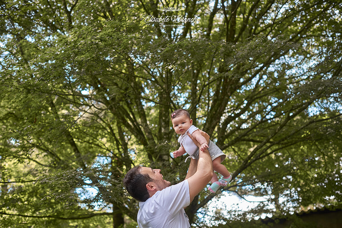 Pai brincando com sua bebê de 4 meses, a levantando alto. Ao fundo os galhos da copa de uma árvore, em Ana Rech, Caxias do Sul.