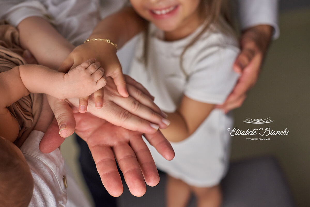 Detalhe das mãos do pai, mãe, filha mais velha e filho caçula, uma sobre a outra, e sorriso da filha mais velha ao fundo, no ensaio newborn em Caxias do Sul.