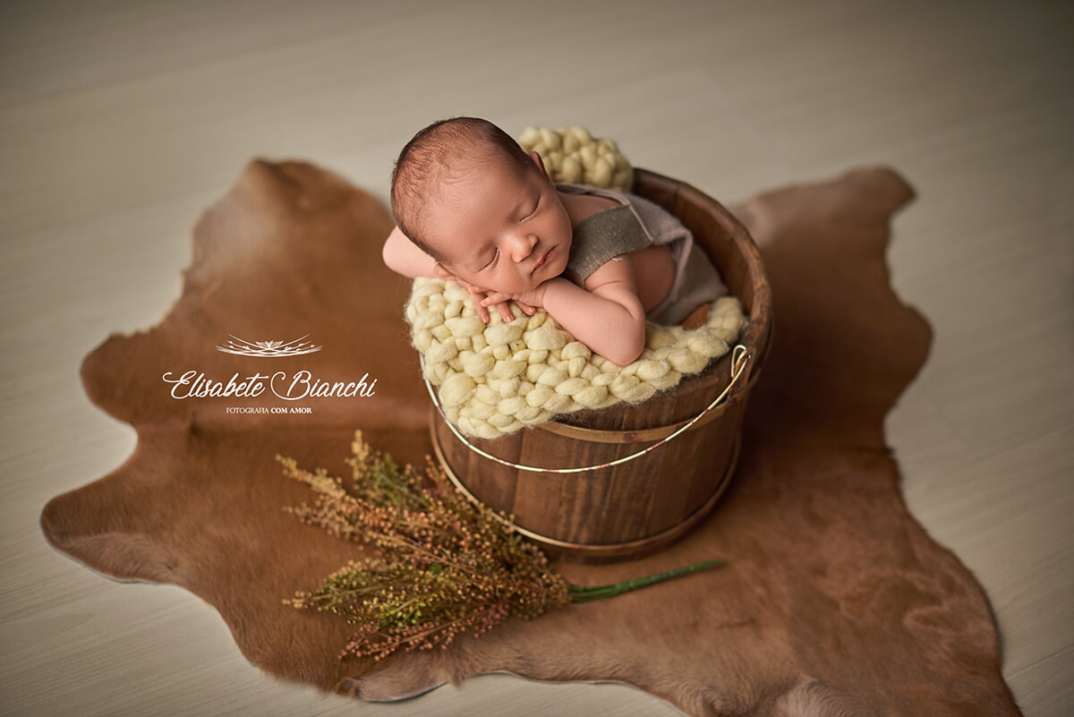 Bebê recém-nascido dormindo  dentro de um baldinho de madeira, no ensaio newborn, em Caxias do Sul.