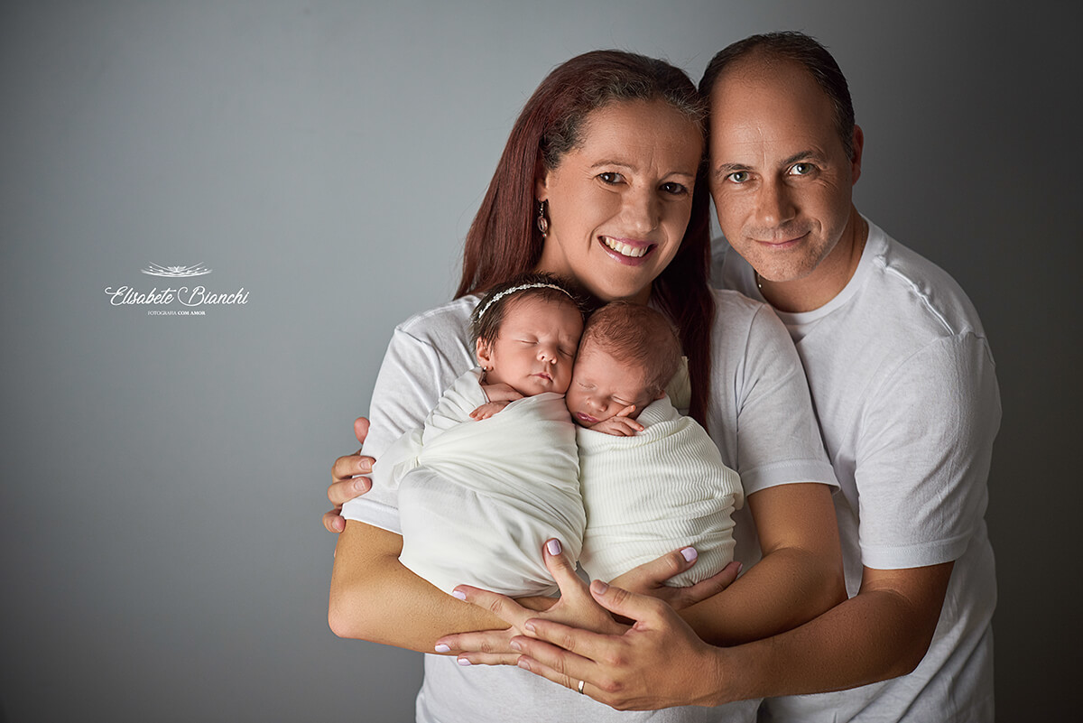 Pais com seu casal de bebês recém-nascidos, gêmeos, no colo da mãe, em ensaio newborn, Caxias do Sul.