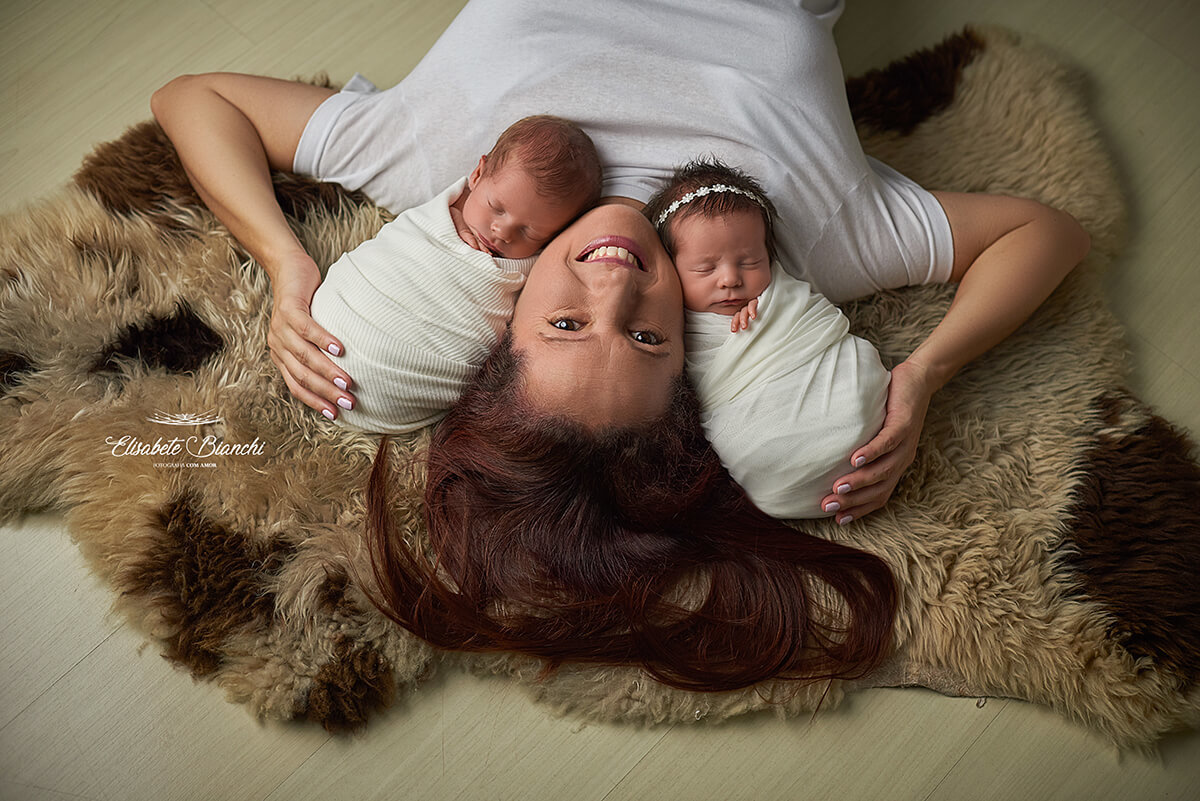 Mãe com seu casal de bebês recém-nascidos, gêmeos, deitados sobre um pelego, em ensaio newborn, Caxias do Sul.