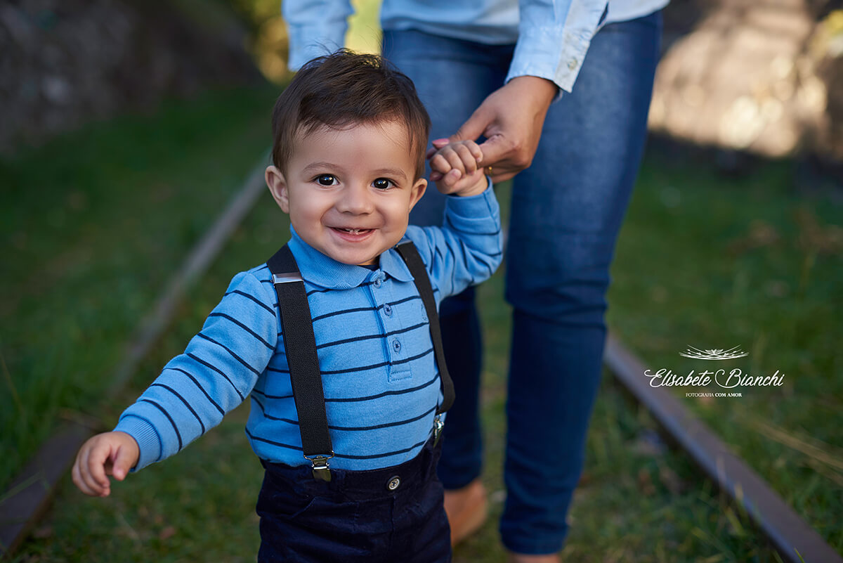 Menino de 10 meses sorrindo e ensaiando os passinhos, segurando o dedo da mãe, em ensaio fotográfico nos trilhos, em Forqueta, Caxias do Sul.