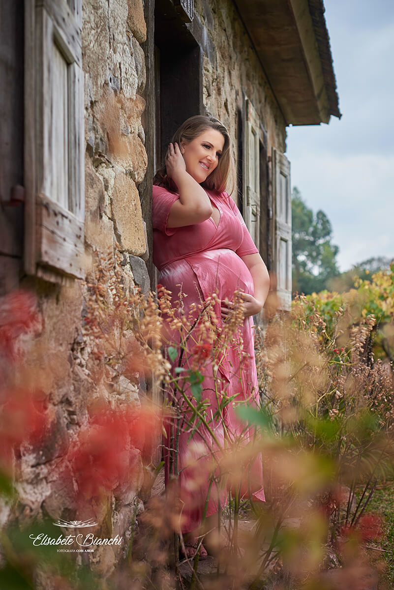 Gestante ajeitando o cabelo, de pé, vista de lado, em frente à uma casa de pedra antiga, em ensaio na Terceira Légua, Caxias do Sul.
