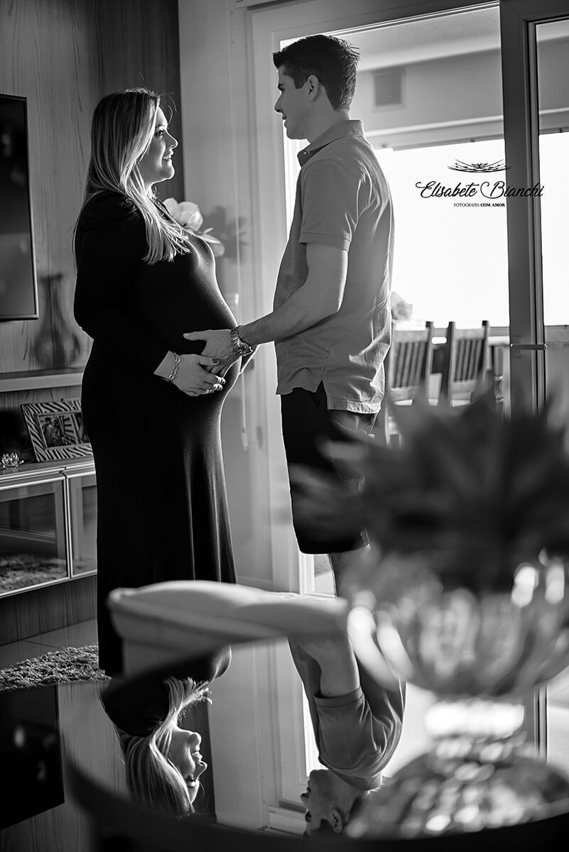 Casal gestante em tempo de pandemia, em casa, de pé em sua sala, olhando um para o outro, com as mãos na barriga. Reflexo do rosto deles na mesa espelhada. Ensaio fotográfico em Caxias do Sul.