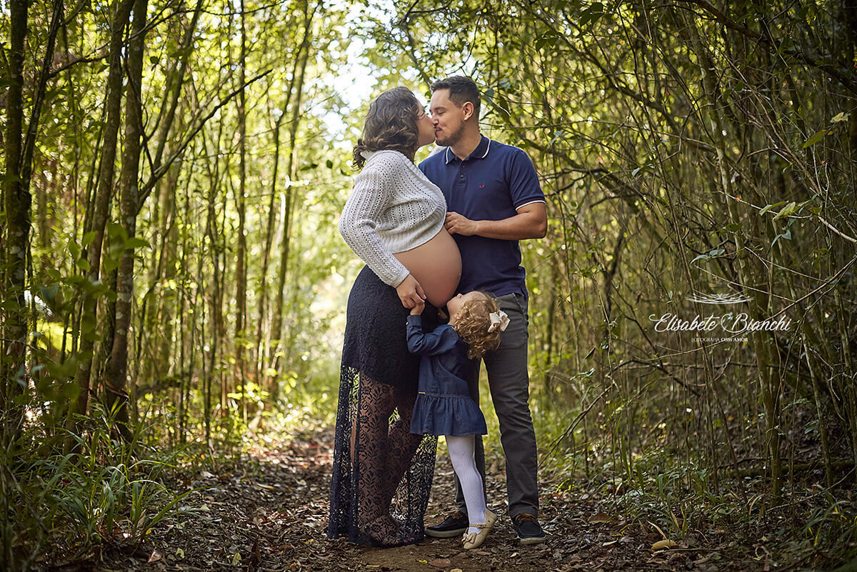 Menina de 2 anos de pontinha dos dedos dos pés, para alcançar a barriga da mãe para beijar seu maninho ainda na barriga,  enquanto sua mãe e seu pai se beijam em um ensaio fotográfico gestante no meio de uma trilha, em Caxias do Sul.
