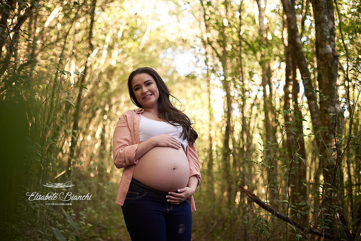 Gestante de frente, abraçando sua barriga exposta, em ensaio da gestação em uma floresta iluminada, em Caxias do Sul.