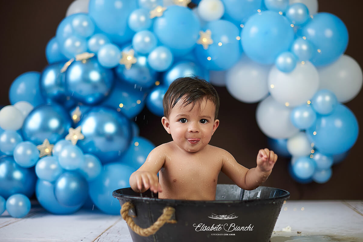Bebê de 1 ano tomando banho na tina, após destruir um bolo no ensaio smash the cake, e, ao fundo, arco de balões azuis desconstruído, em ensaio fotográfico em Caxias do Sul. 