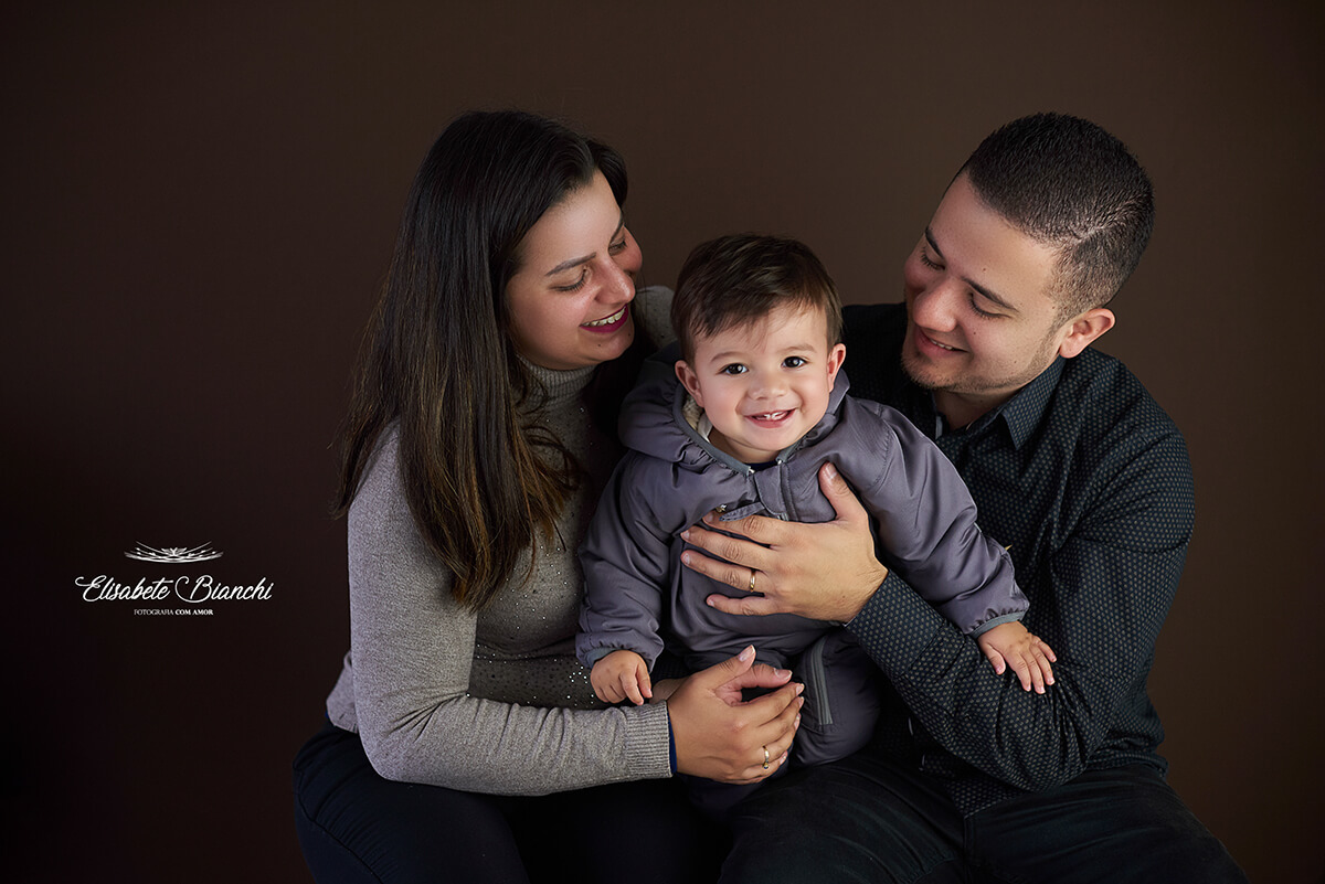 Pais segurando seu filho de 1 ano de idade, sobre fundo marrom, em Caxias do Sul.