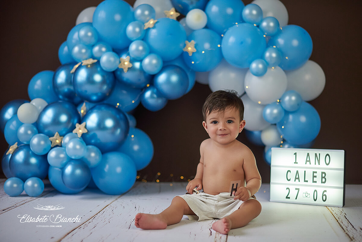 Bebê com 1 ano de idade sentado e sorrindo. Ao fundo, um arco de balões azuis desconstruído. Ensaio fotográfico em Caxias do Sul.