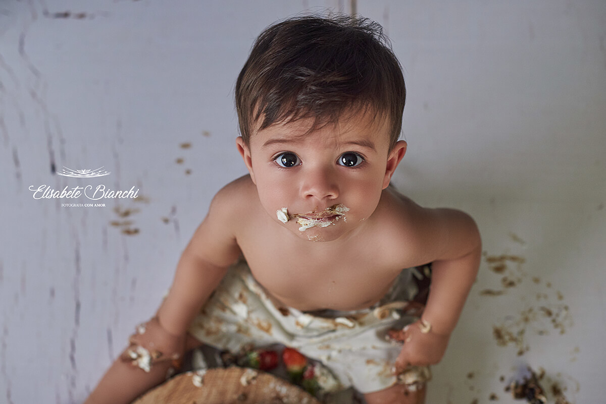 Bebê melecado de bolo, olhando para a câmera, visto de cima para baixo, em ensaio fotográfico smash the cake, em Caxias do Sul.