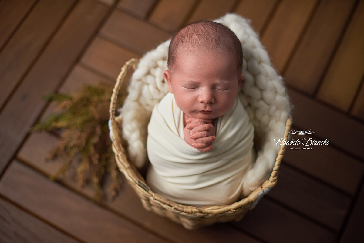 Bebê recém-nascido com as mãos em oração, enroladinho e dentro de um cesto, no ensaio newborn em Caxias do Sul.