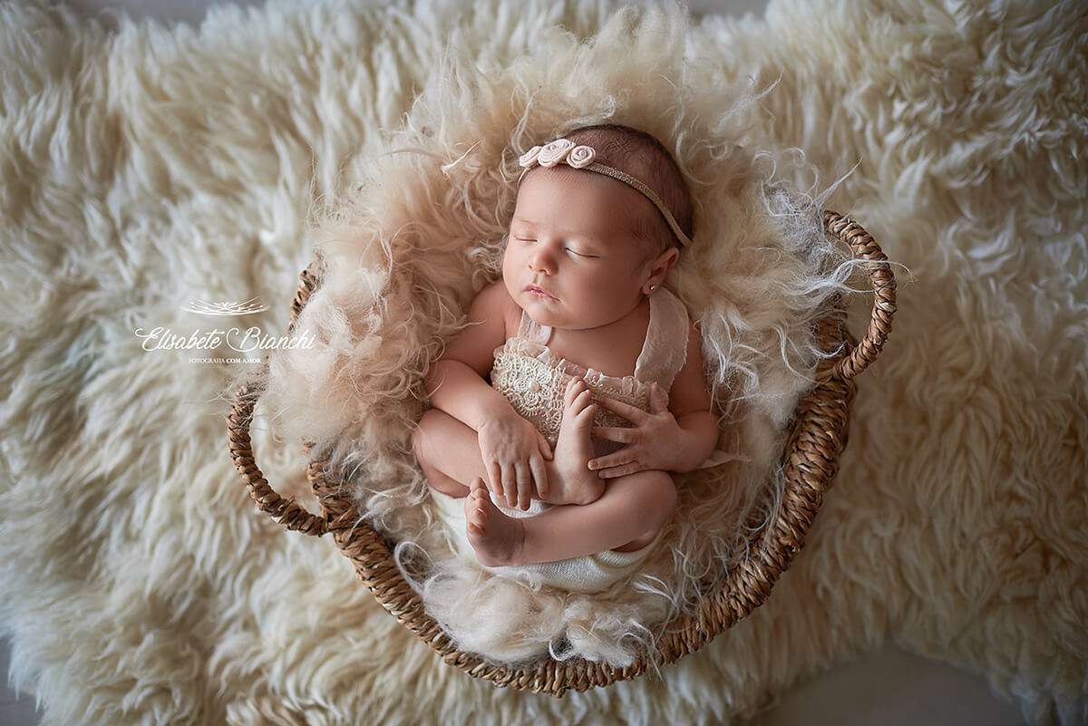 Bebê recém-nascida, deitada de barriga para cima, segurando o próprio pé, deitada em um cesto redondo com pelego, em ensaio newborn, em Caxias do Sul-RS.