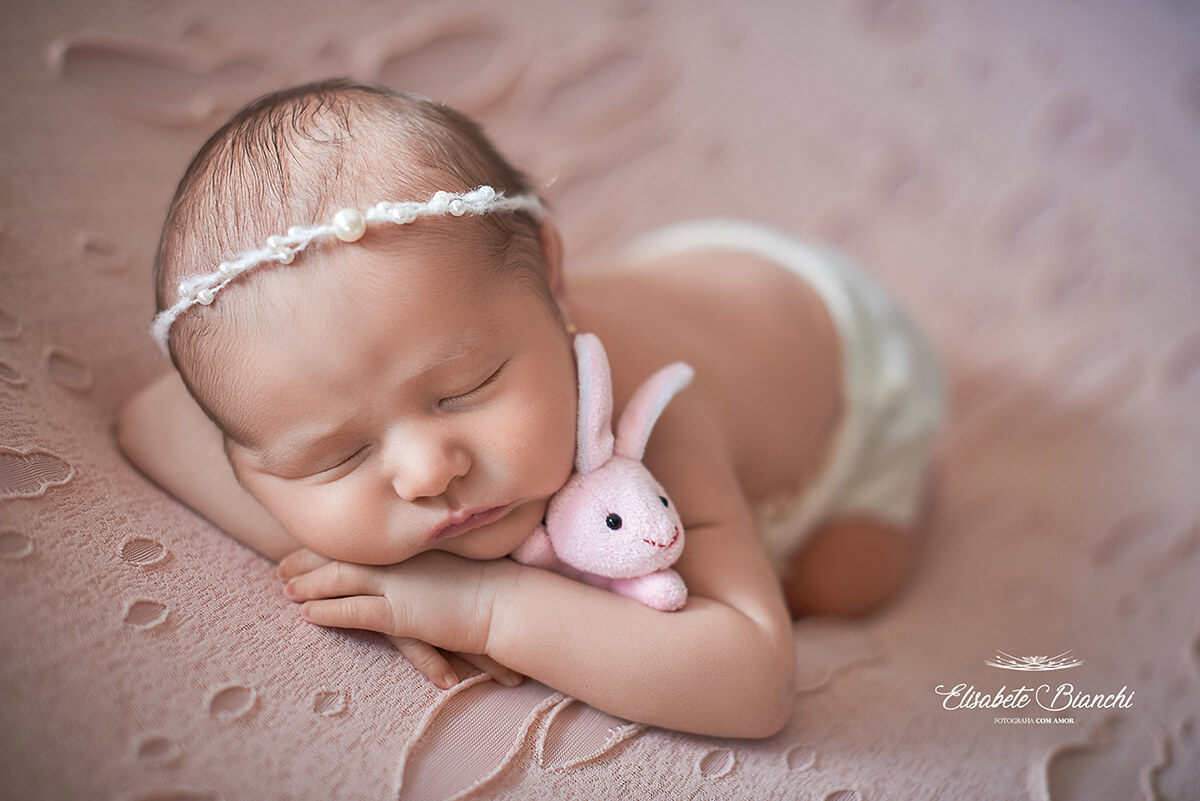 Bebê recém-nascida deitada de bruços, com a cabeça apoiada sobre seus bracinhos e agarrada em um coelhinho miniatura de pelúcia, cor de rosa, em ensaio newborn, em Caxias do Sul-RS.
