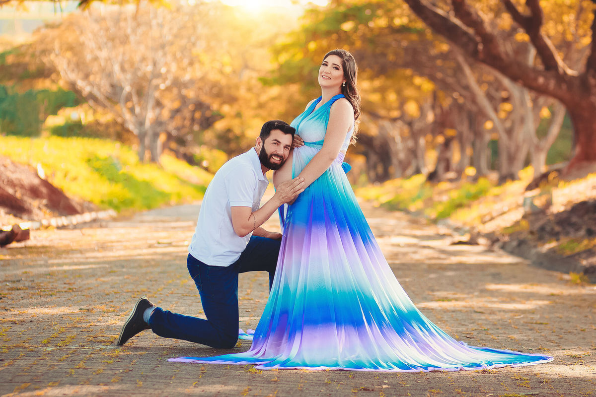 Papai ajoelhado beijando a barriga da mamãe que está com vestido arco íris, no caminho dos flamboyant, em santa bárbara d'oeste - SP