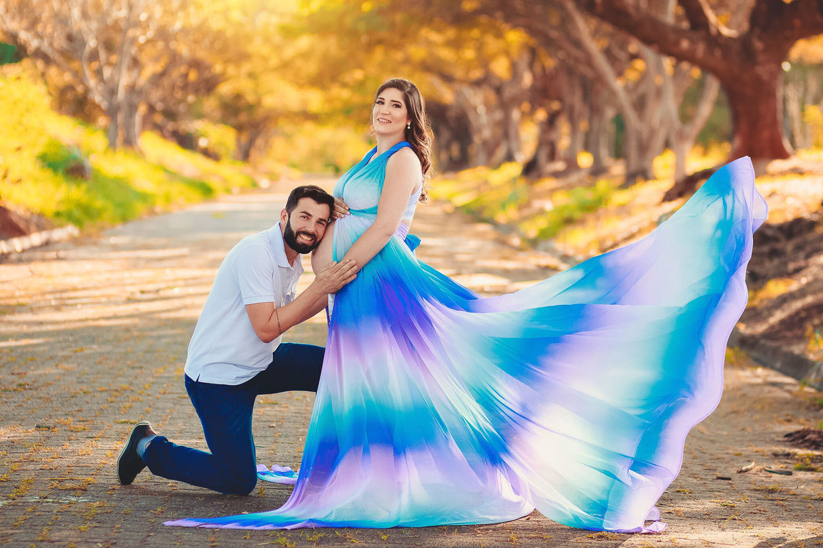 Papai ajoelhado beijando a barriga da mamãe que está com vestido arco íris, no caminho dos flamboyant, em santa bárbara d'oeste - SP