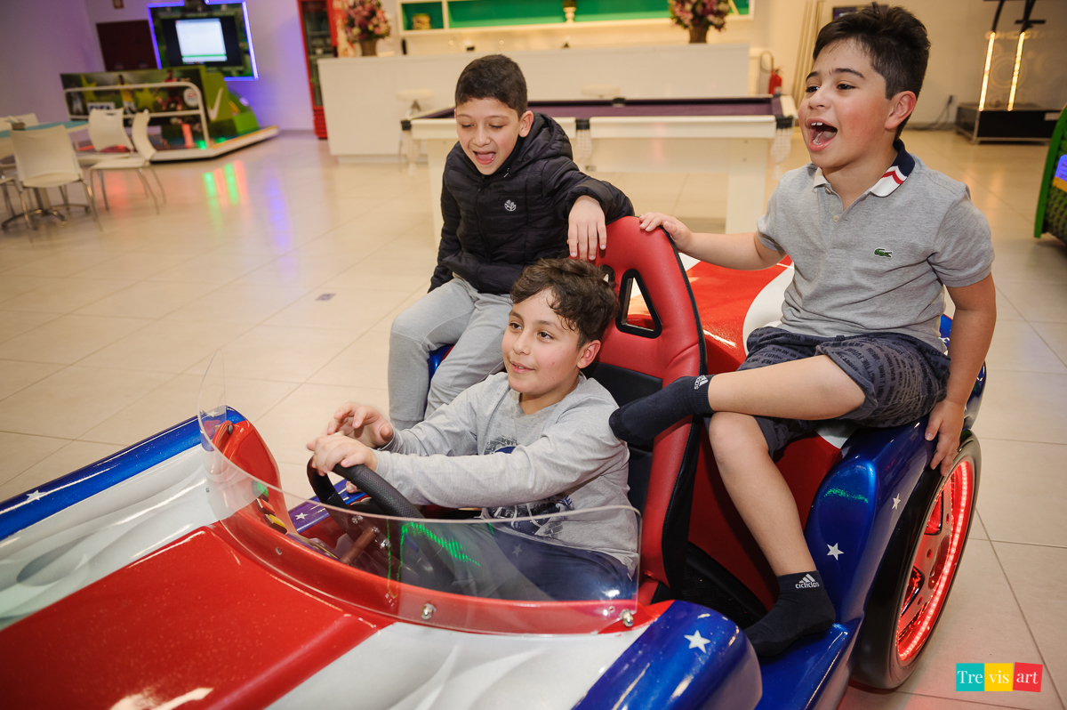 Foto aniversariante brincando no simulador de carro com amigos no buffet de festa infantil.
