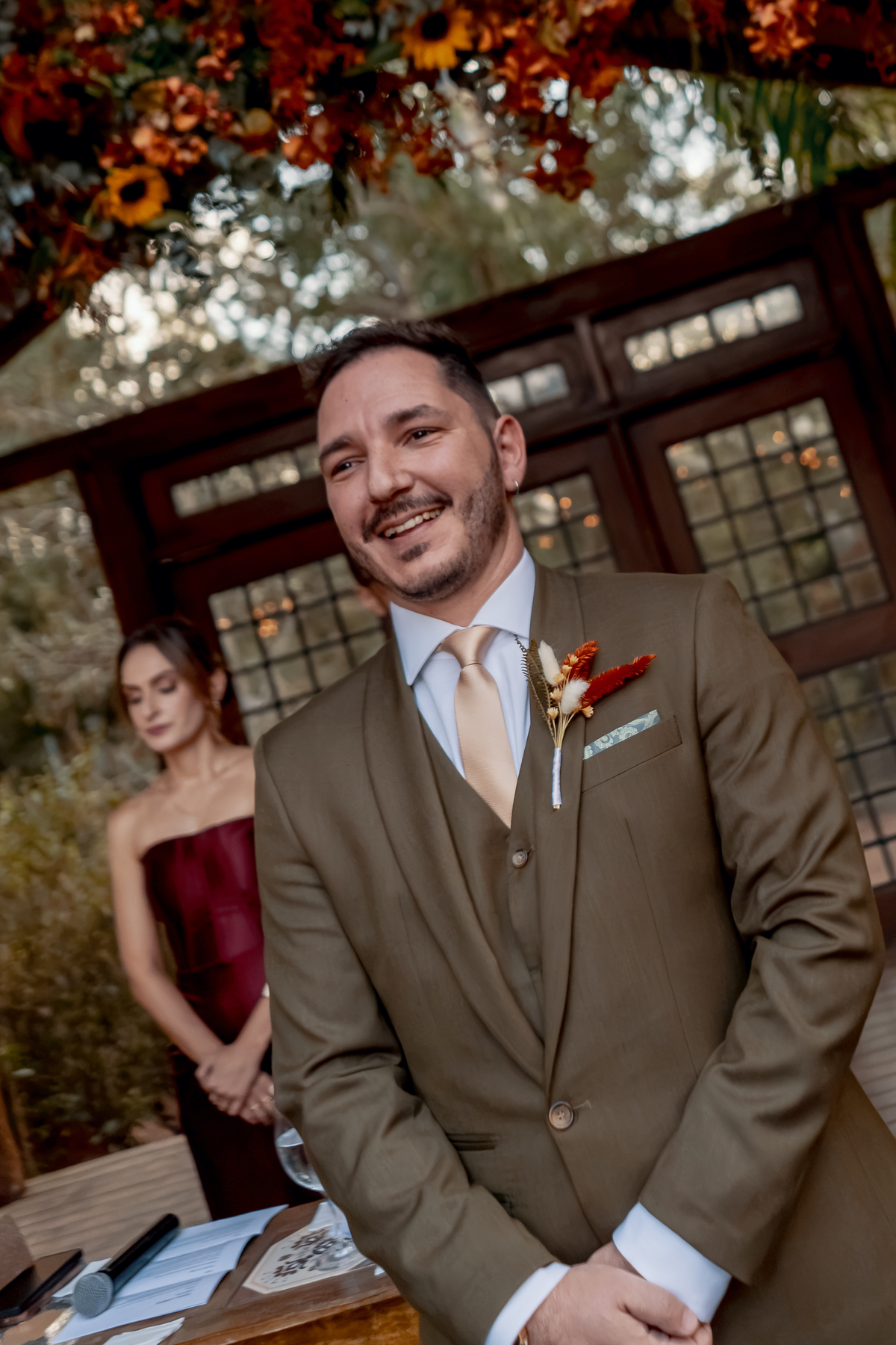 Fábio sorrindo durante a cerimônia, com Yasmin ao fundo e os convidados acompanhando o momento do casamento na Fazenda Vila dos Eucaliptos.