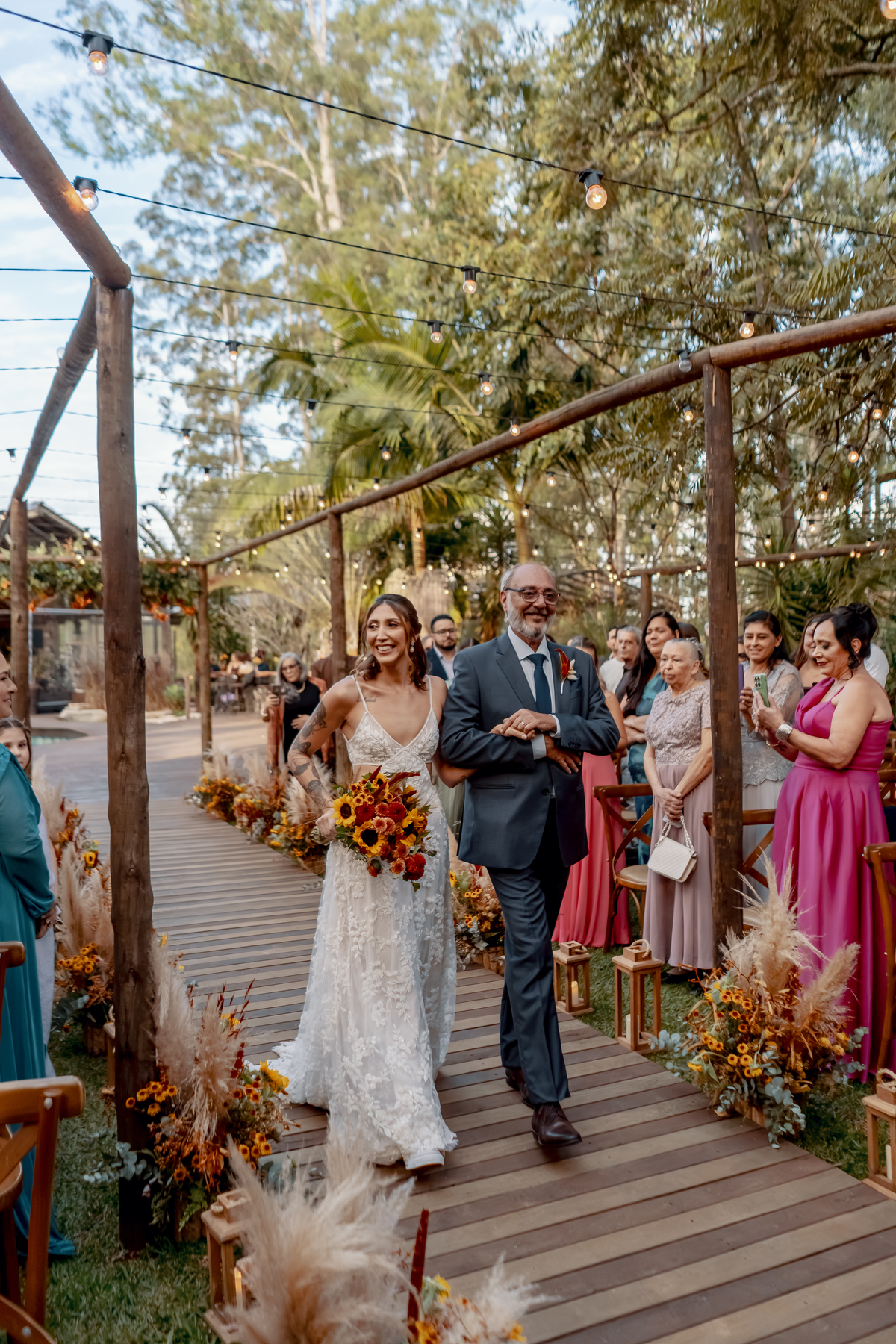 Yasmin caminhando em direção ao altar ao lado de seu pai, cercada pelos convidados e pela decoração floral do casamento ao ar livre.