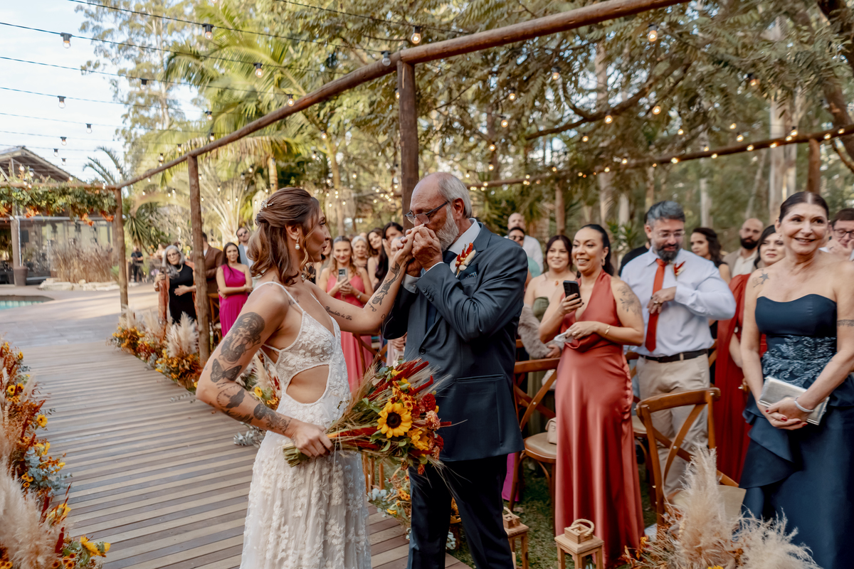 Entrada de Yasmin para a cerimônia de casamento, acompanhada pelo pai e recebida pelos familiares e amigos em meio à natureza.