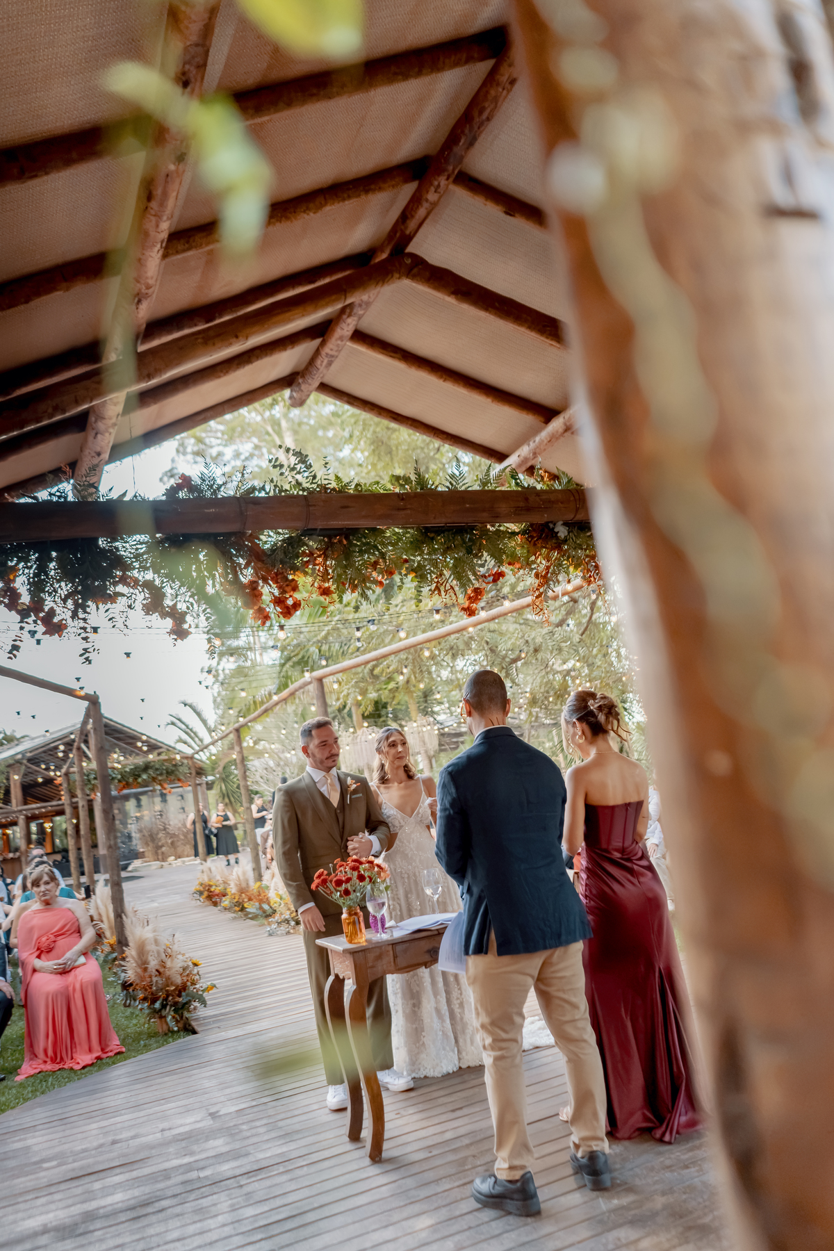 Momento da cerimônia visto através da decoração, com Fábio e Yasmin ao fundo e a arquitetura rústica da fazenda compondo a fotografia.
