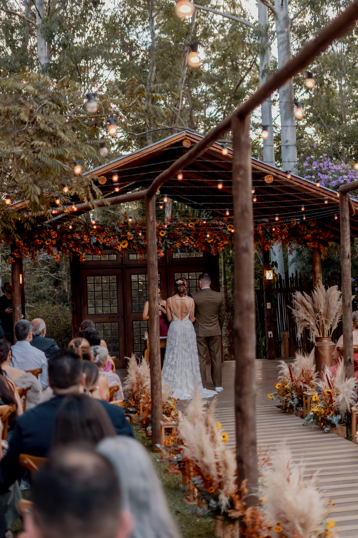 Yasmin entrando para a cerimônia de casamento na Fazenda Vila dos Eucaliptos, sob a estrutura rústica decorada com flores e folhagens.