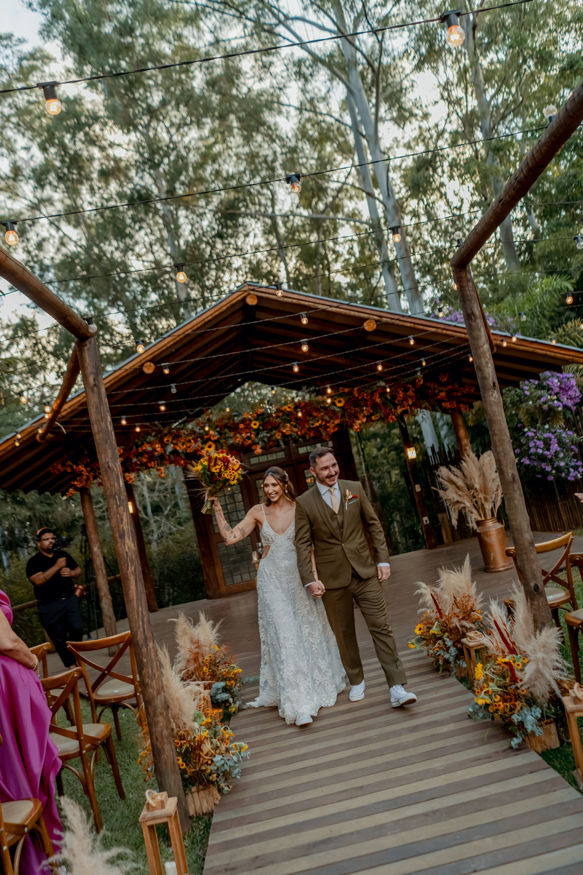Fábio e Yasmin diante do altar da Fazenda Vila dos Eucaliptos, com a decoração floral e a estrutura rústica ao fundo.
