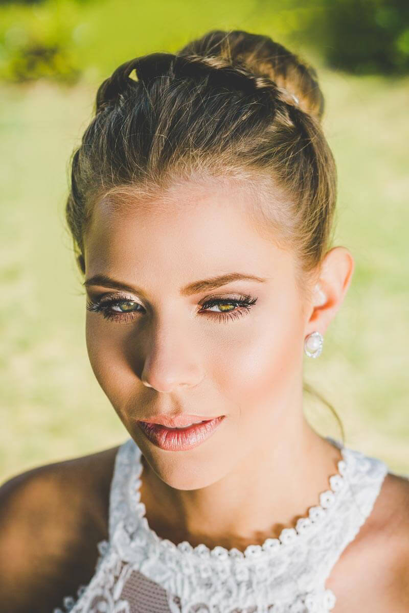 Noiva maquiada para casamento ao ar livre ou para igreja de dia. Com cabelo preso tipo coque e usando joias