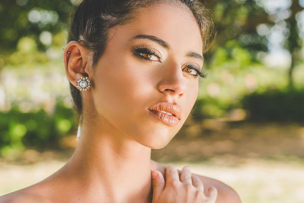 Noiva maquiada para casamento ao ar livre ou para igreja de dia. Com cabelo preso tipo coque e usando joias