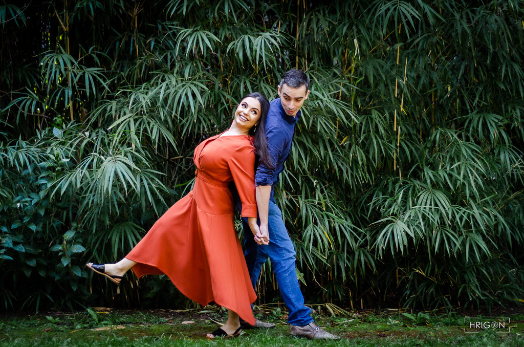casal de costas um para o outro dando as mãos. ela está se jogando um pouco em cima das costas dele de forma divertida. ao fundo árvores verdes de bambu