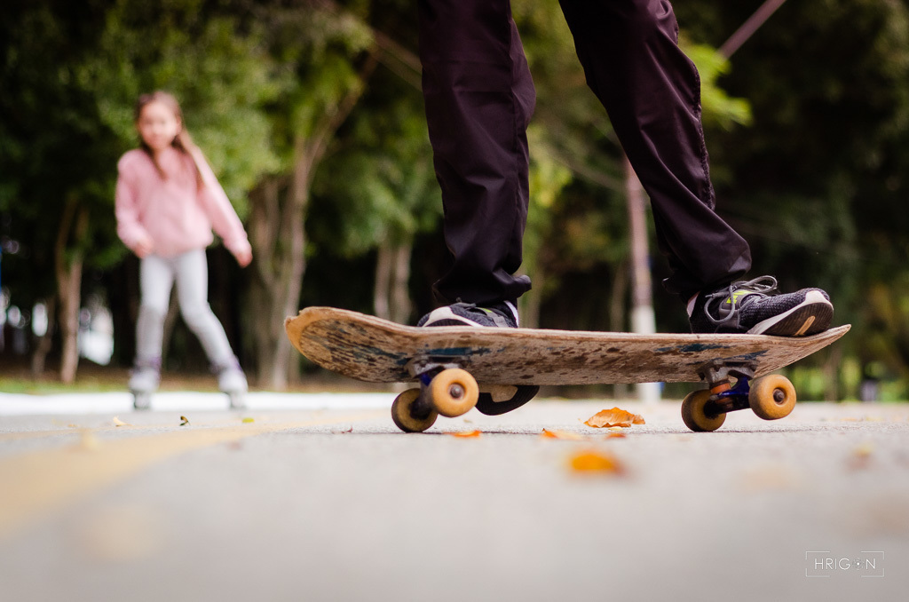 em primeiro plano temos o skate no asfalto enquanto a filha do casal se aproxima de patins, ela está bem desfocada ao fundo