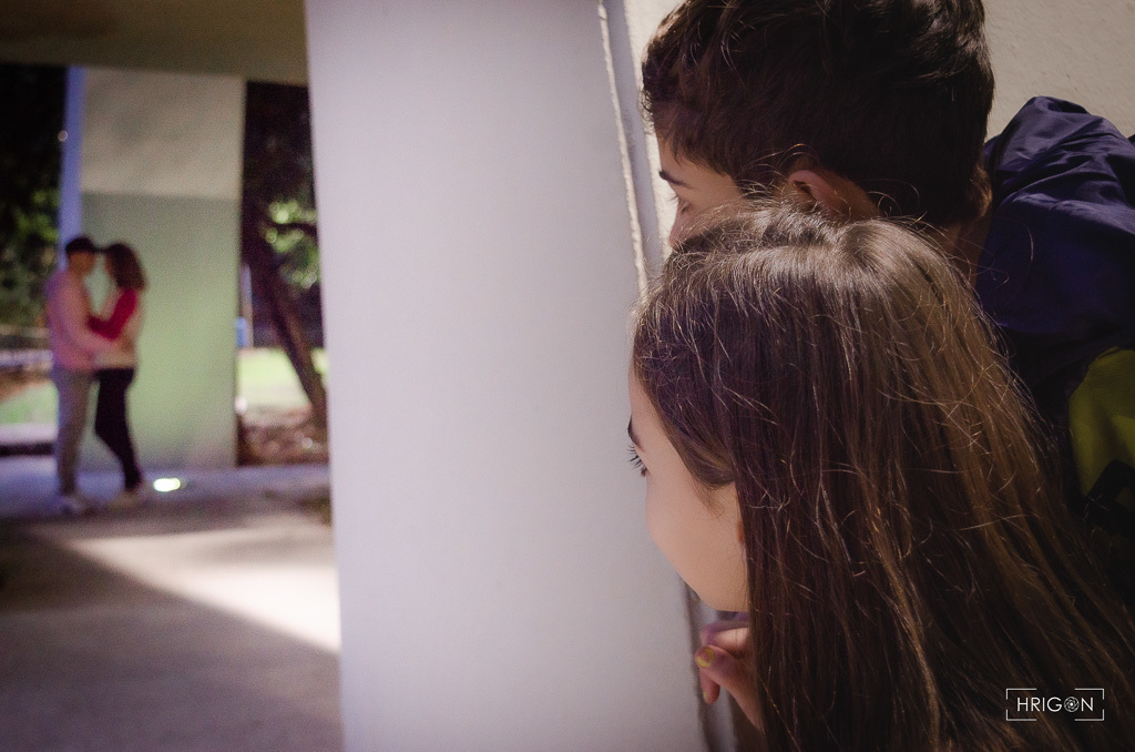 os pais estão frente a frente quase se beijando embaixo de um monumento do parque, enquanto os filhos estão espiando atrás do muro