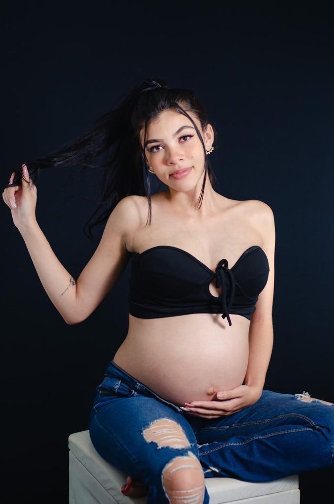 Foto gestante top preto calça jeans rabo de cavalo fundo preto sentada no puff 