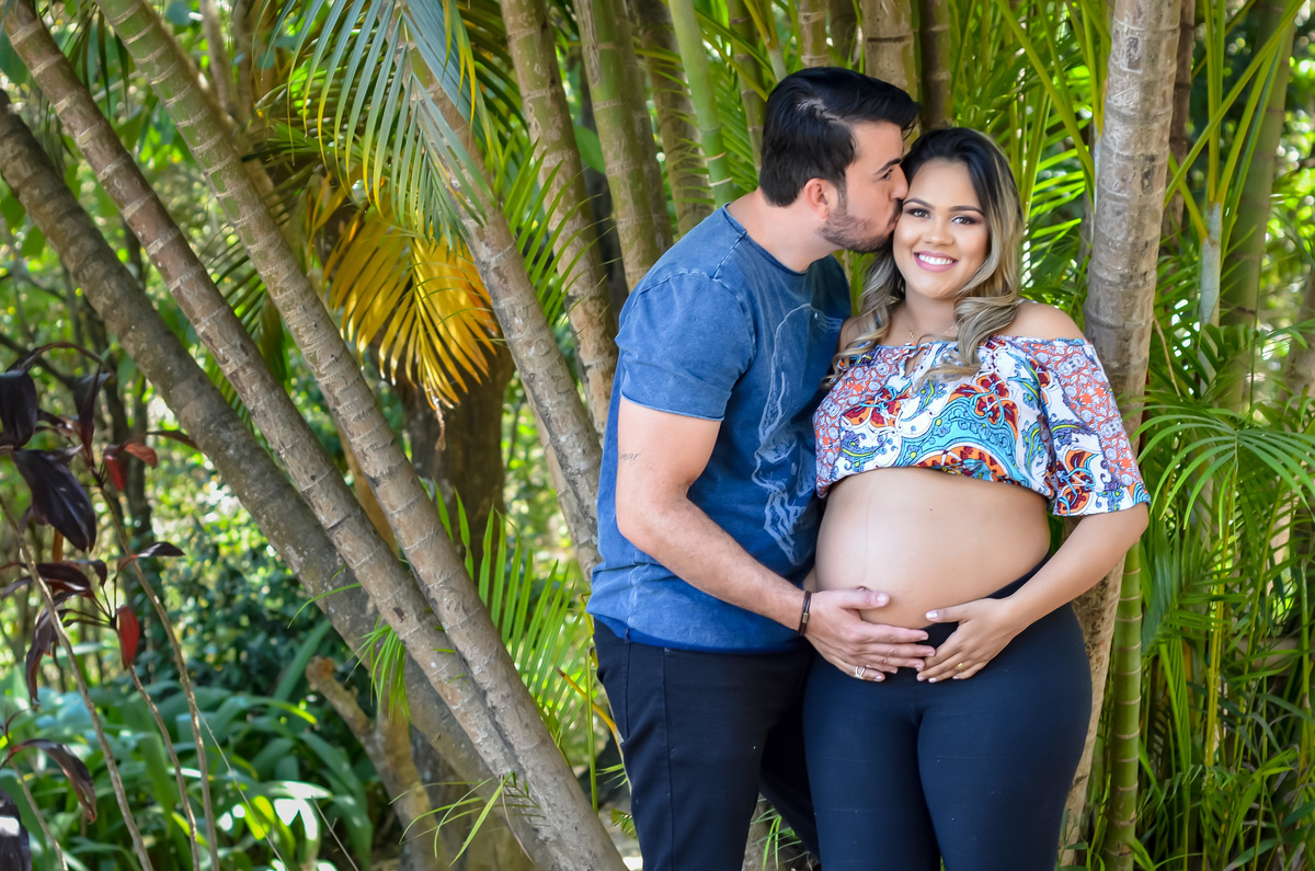 Casal gestante se beijando em meio a árvores e muito verde Tendo como plano de fundo um coqueiro dê jardim 