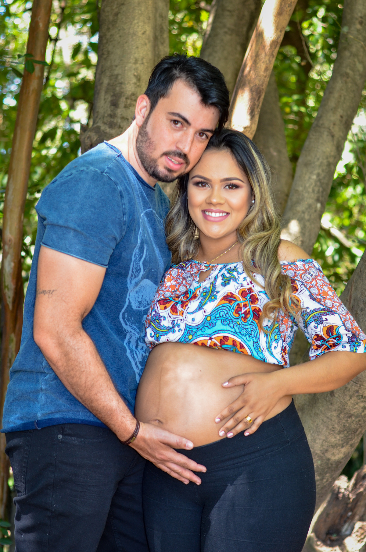 Foto casal gestante abraçados sorrindo em um dia ensolarado em meio a árvores 